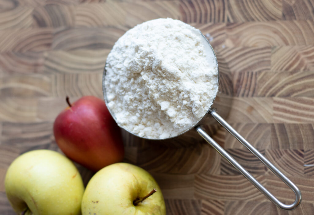 measuring cup with flour and several apples on a wooden board.